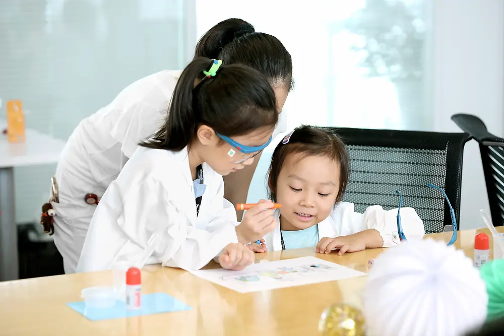 Dvoje djece i žena u laboratorijskoj kuti boje sliku za stolom