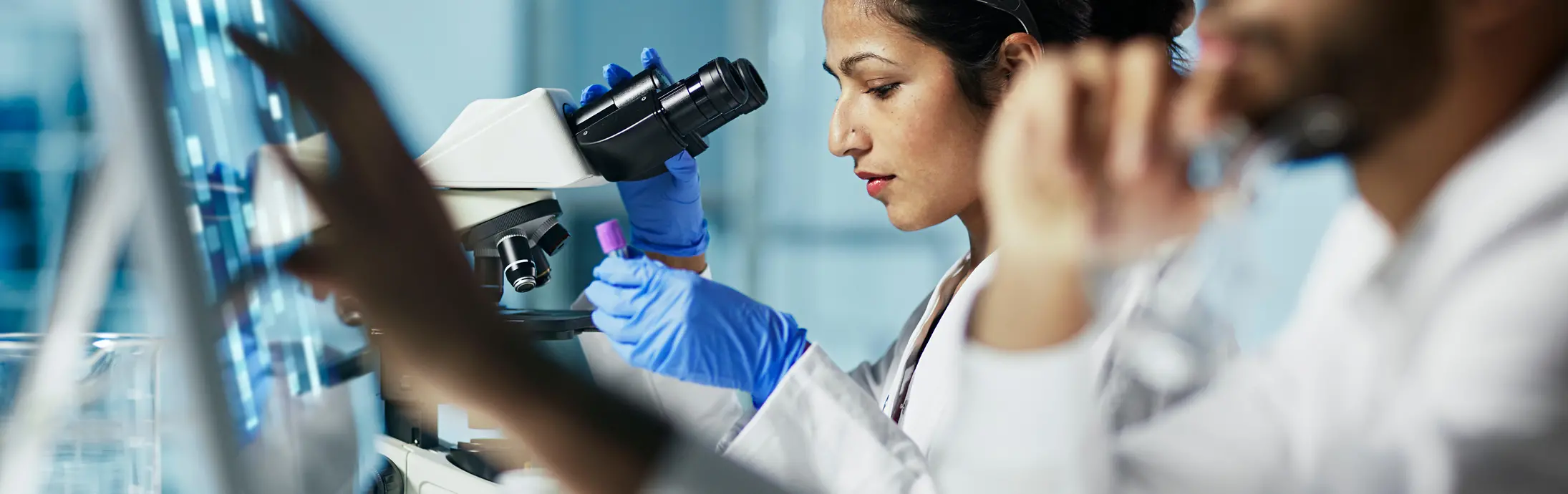 žena koja u laboratoriju sjedi i gleda uzorak kroz mikroskop; ispred nje je muškarac s bradom koji gleda u zaslon 