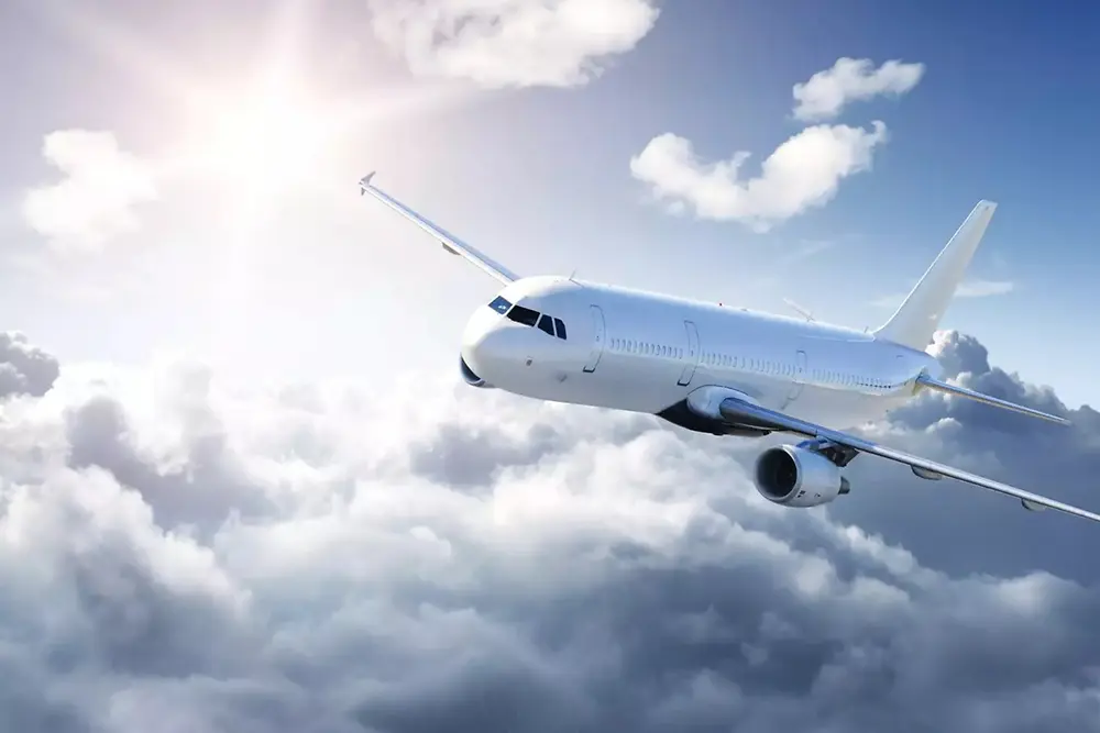Bijeli putnički avion leti iznad mekanih oblaka na svijetlom nebu, uz sunčevu svjetlost iz gornjeg lijevog kuta.
