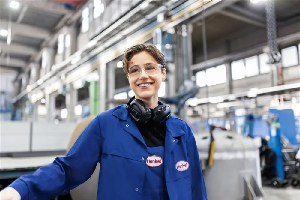 Nasmijana zaposlenica Henkela u plavom radnom kaputu i sa zaštitnim slušalicama stoji u industrijskoj proizvodnoj hali, predstavljajući stručnost i inovacije u proizvodnji.