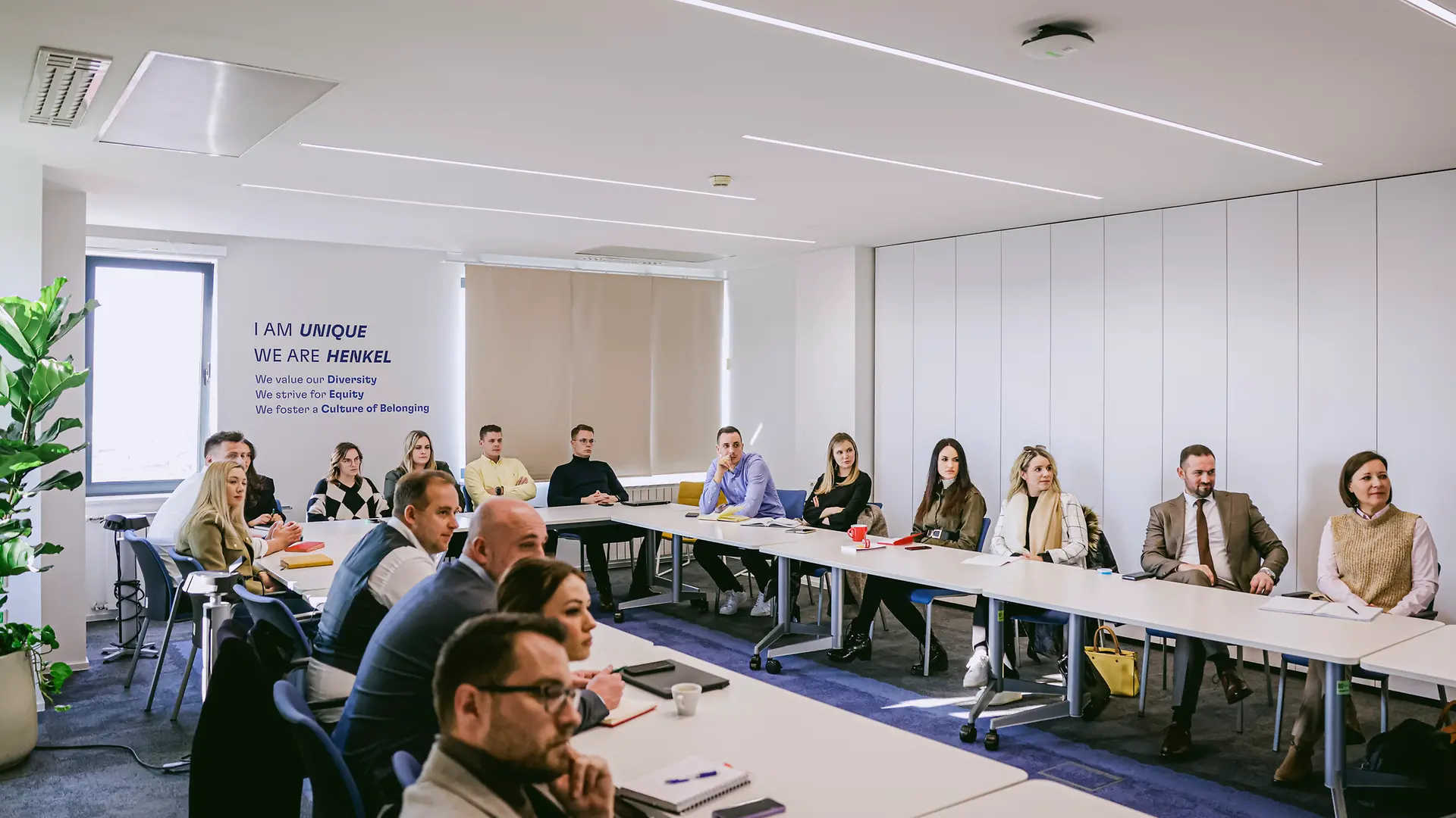 Na fotografiji je konferencijska dvorana s dugim stolovima postavljenim u obliku slova U, u kojoj više osoba sudjeluje na sastanku.