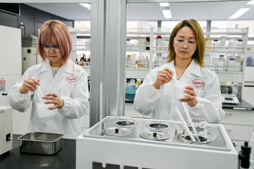 Dvije osobe u bijelim Henkelovim laboratorijskim ogrtačima rade u modernom laboratoriju. Rukuju znanstvenim instrumentima i spremnicima na radnoj površini, a u pozadini su vidljiva različita laboratorijska oprema i police s potrepštinama.