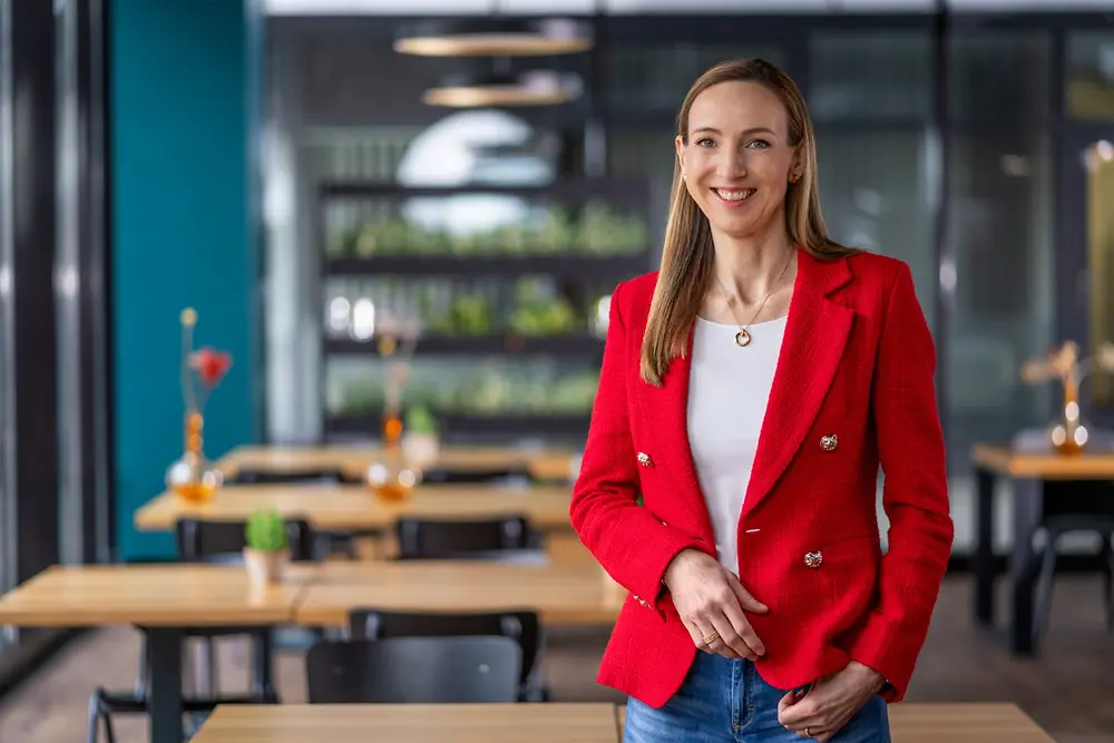 Dr. Simone Bagel-Trah wearing a bright red blazer and white top standing in a modern cafeteria environment with wooden tables, and plants in the background.