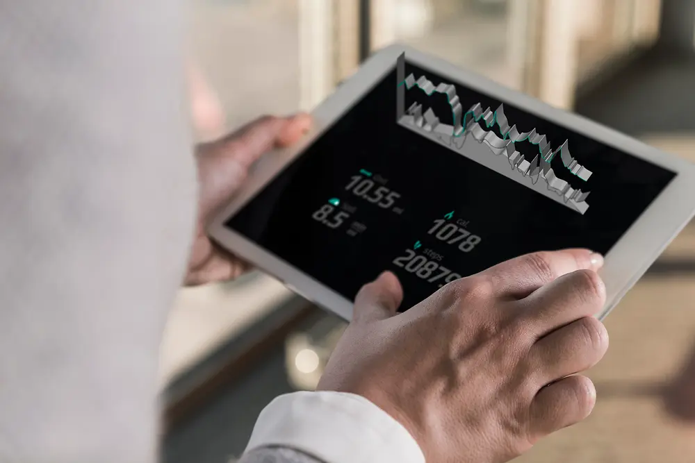 Osoba drži tablet na kojem su prikazani digitalni grafikoni i numerički podaci, gledano preko ramena u svijetlom zatvorenom prostoru.